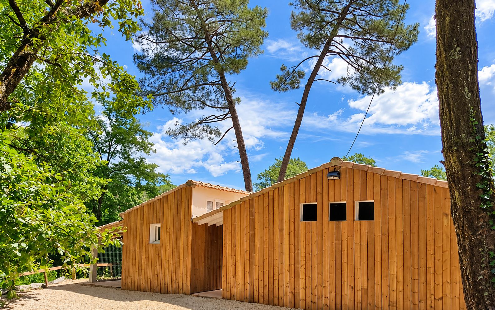 Un bâtiment en bois à bardage vertical et toit de tuiles, entouré de pins et de feuillages sous un ciel bleu nuageux.