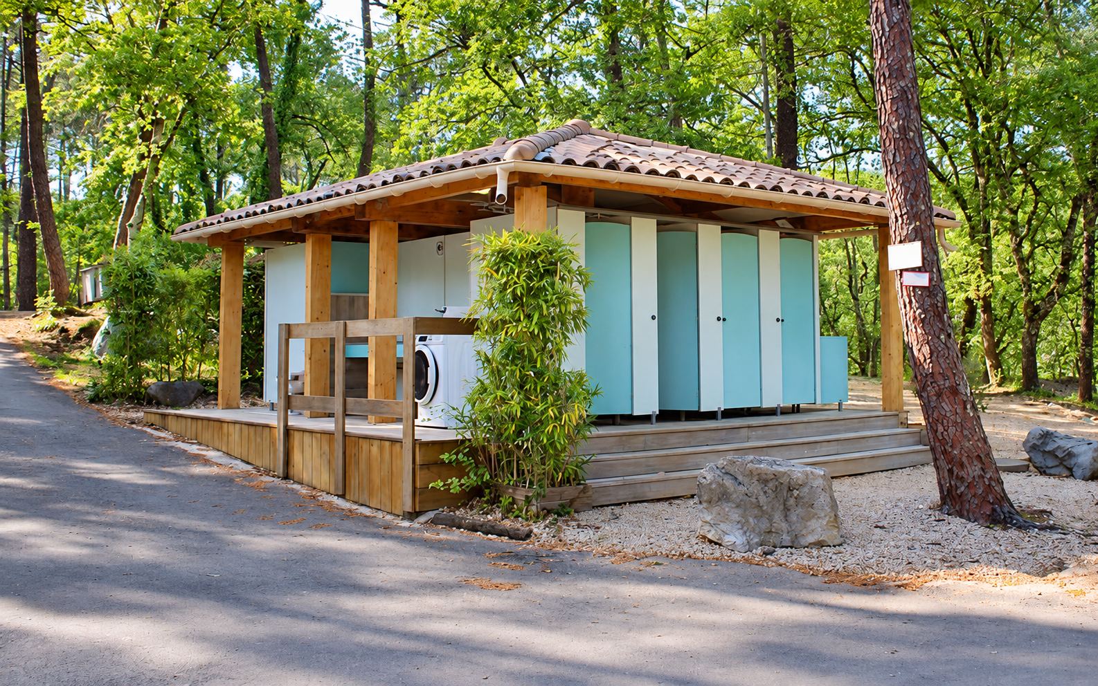 Un bloc sanitaire en bois au toit de tuiles, avec des cabines turquoise et une machine à laver, entouré de pins.