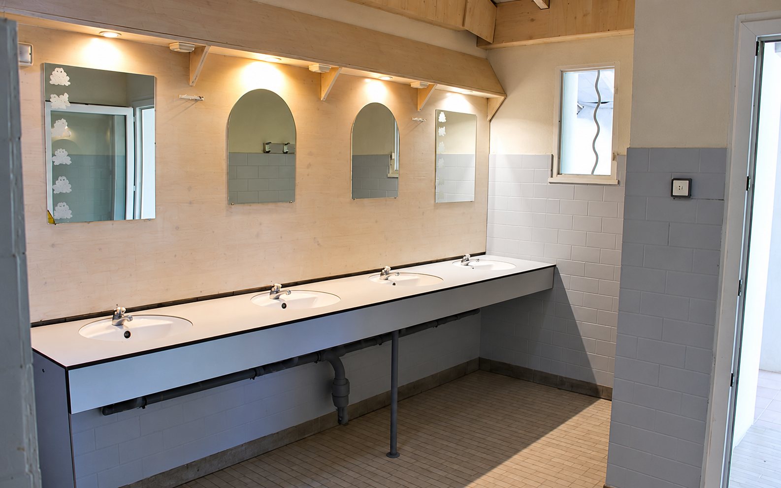 Salle d'eau commune avec une longue rangée de lavabos blancs, des miroirs au mur carrelé et des spots lumineux au plafond.