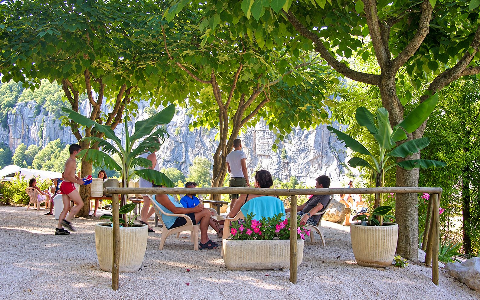 Des vacanciers se détendent sur des chaises en bois à l'ombre de grands arbres, entre des bacs de fleurs, devant une falaise calcaire.